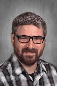 Close-up of a smiling man wearing black glasses and a plaid shirt, neutral gray backdrop.
