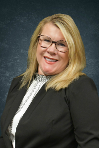 A smiling blonde woman wearing tortoiseshell glasses and a black blazer, posing for a professional headshot against a textured dark backdrop.